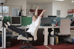 Side view of cheerful female employee in formal outfit sitting on office chair with crossed legs on desk and stretching while resting during work with closed eyes