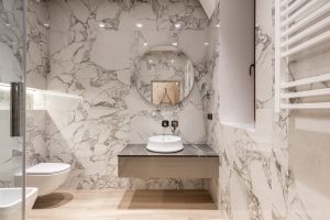 Interior of modern bathroom with round mirror hanging on marble wall and white ceramic sink and toilet bowl